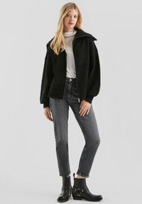 Zwarte donzige jas met een hoge kraag; lange mouwen. Gepaard met grijze jeans en enkellaarzen met gespsaccenten. Eenvoudig, modern ontwerp.