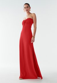 Lange rode strapless jurk met een gedraaide corselet, een zijsplit en een gladde stofstructuur. Geaccentueerd met eenvoudige metalen sieraden.