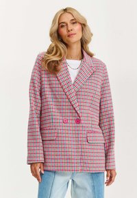 Žena s blond vlasy, která má na sobě barevnou sako s růžovými, modrými a oranžovými kostkami a světle modré džíny, stojící na jednobarevném pozadí.