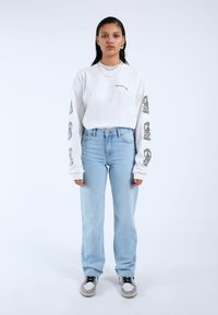 Camisa blanca de manga larga con diseños gráficos negros en las mangas, combinada con jeans de mezclilla azul claro y zapatillas gris con cordones negros.