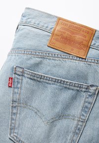 Vaqueros de mezclilla azul claro con un parche trasero de cuero que presenta texto y gráficos en relieve. Pestaña roja de Levi's en el bolsillo trasero derecho.
