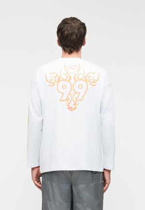 Férfi fehér hosszú ujjú ingben, narancssárga díszítő lángokkal és a "99" szám hátsó részén, szürke nadrággal, háttal nézve.