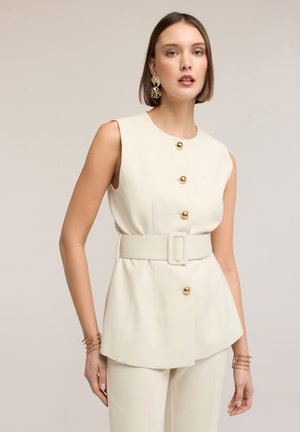 Kvinde iført ærmeløs cremefarvet blazer med bredt bælte og guldknapper, sat sammen med matchende bukser samt store guldøreringe og armbånd.