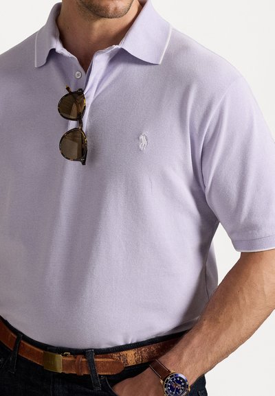 Ανοικτό μοβ πόλο μπλούζα με υφή υφάσματος, διαθέτοντας ένα μικρό κεντημένο λογότυπο, φορεμένη με γυαλιά ηλίου και καφέ δερμάτινη ζώνη.