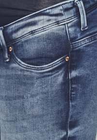 Vaqueros de mezclilla en azul oscuro con un bolsillo frontal, un ligero desgaste y detalles de remaches de cobre en el bolsillo. Tejido suave y texturizado.