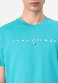 Turquoise katoenen T-shirt met het "TOMMYJEANS" logo in het wit, met daaronder een klein rood, wit en blauw embleem. Klassiek ontwerp met ronde hals.