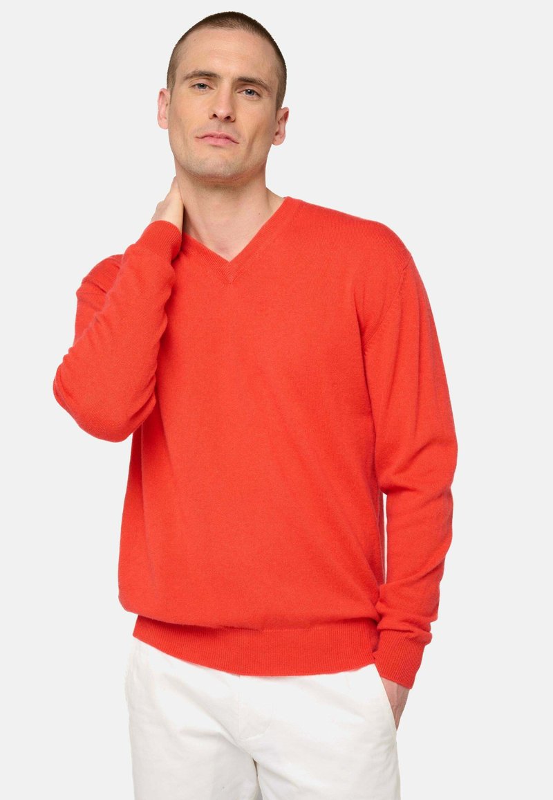 Pull à col en V rouge en texture tricotée, avec des manches longues et des poignets côtelés. Associé à un pantalon blanc, affichant une silhouette décontractée.