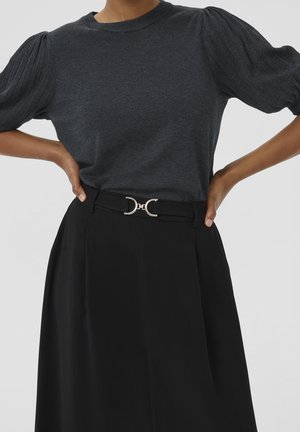 Femme portant un haut tricoté gris foncé à manches bouffantes, rentré dans une jupe taille haute noire avec une boucle de ceinture décorative argentée.