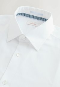 Weiße Hemdbluse mit glatter Textur, Knopfverschluss und einem Kragen mit innenliegender Navy-Musterung. Das Etikett trägt die Aufschrift "NEXT SIGNATURE."