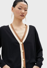 Black cardigan with a V-neck, contrasting brown and cream trim, and gold buttons. Features loose-fitting sleeves and textured fabric.