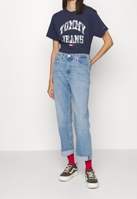 Persona vistiendo una camiseta de Tommy Jeans de color azul marino, jeans claros con dobladillo, calcetines rojos y zapatillas con estampado, de pie contra un fondo blanco liso.