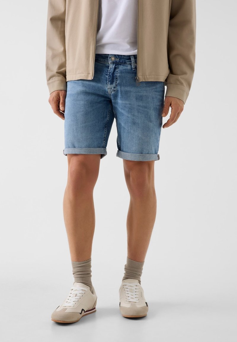 Man met opgerolde lichtblauwe denim shorts, beige jas, wit T-shirt, beige sokken en witte sneakers, staand tegen een grijze achtergrond.