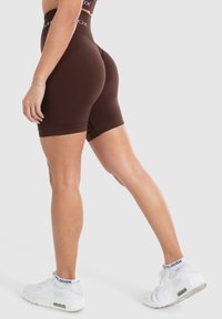 Shorts corti marroni a vita alta realizzati in materiale elastico, con una texture liscia e cuciture sottili, abbinati a sneakers bianche da ginnastica.