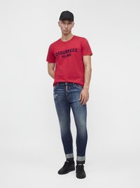 Homme portant un t-shirt rouge DSQUARED2 Milano, un jean bleu délavé avec les revers roulés, des baskets noires et une casquette baseball noire, debout devant un fond blanc.