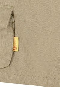 Tessuto beige con una trama texture, caratterizzato da una tasca cucita e un'etichetta gialla prominente con logo rosso. Dettagli di design semplici e funzionali.