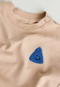 Világos rózsaszín anyagú póló, amelyen egy kék kötött háromszög alakú folt látható, rajta egy mosolygós arc és két patentgomb a vállnál.