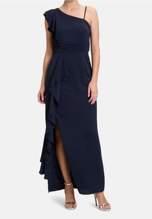 Vrouw draagt een marineblauwe avondjurk met één schouder, met ruches en een zijsplit, gecombineerd met zilveren sandalen met hoge hak.