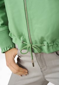 Grüne Leder-Bomberjacke mit Kordelzug in der Taille, gerippten Bündchen und metallischen Reißverschlüssen; über hellgrauten Hosen getragen.
