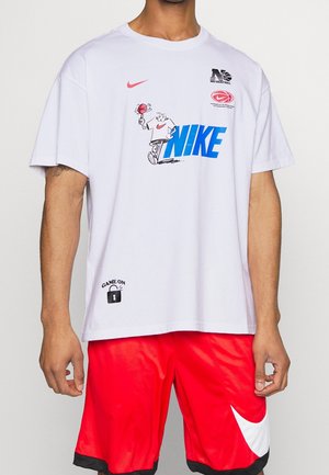 Osoba nosząca białą koszulkę Nike z grafiką i logo związanymi z koszykówką, w połączeniu z czerwonymi spodenkiami do koszykówki Nike z białym znakiem Swoosh.