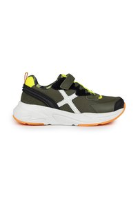 Zapatilla deportiva verde y negra con un diseño de cruz blanca, acentos amarillos, cinta de velcro y suela naranja. Presenta paneles texturizados.