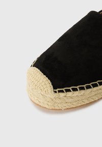 Espadrille en daim noir avec une semelle en jute beige tissée et une bordure noire cousue, présentant un bout arrondi et une tige texturée.