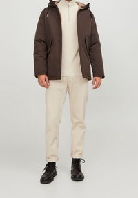 Brun vadderad jacka med huva, dragkedja vid kragen över en beige tröja, kombinerad med ljusa byxor och svarta snörkängor.