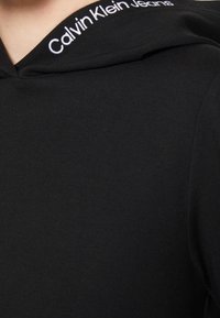 Escote de prenda negra con logotipo "Calvin Klein Jeans" bordado en blanco sobre tela, primer plano.