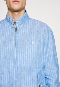 Man som bär en ljusblå linnekavaj med vita vertikala ränder, dragkedja framtill, knappar på kragen och en vit broderad logotyp på bröstet.