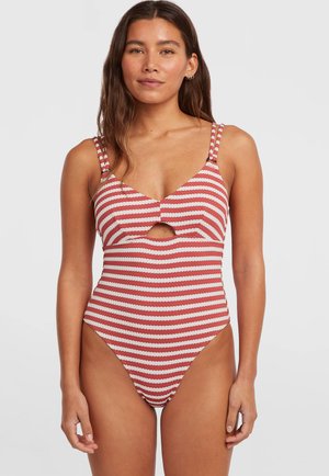 Vrouw die een rood-wit gestreepte eendelige badpak draagt met een voorste uitsnijding en dubbele schouderbandjes, staand tegen een effen achtergrond.