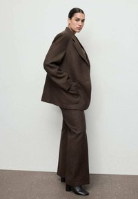 Completo in lana marrone con giacca ampia dotata di rever e pantaloni a gamba larga. Stivaletti neri lucidi con tacco completano il look.