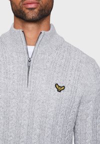 Pull en maille gris avec col montant et fermeture éclair. Présente un écusson brodé jaune et noir sur la poitrine, motif torsadé texturé.