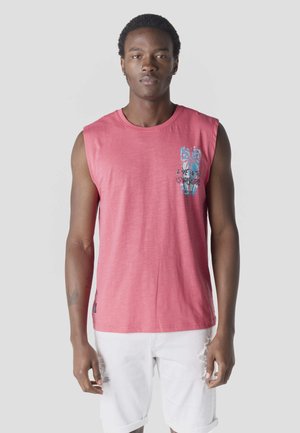 Joven vestido con una camiseta sin mangas roja con estampado azul de palmeras y pantalones cortos blancos rasgados, de pie contra un fondo claro y liso.