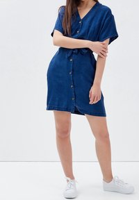 Džínová šaty v tmavě modré barvě. Mají krátké rukávy, véčkový výstřih, zapínání na knoflíky a pásek na zavázání. Nosené s bílými teniskami. Hladká textura.