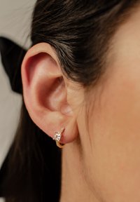 Pendiente de aro dorado con una piedra rectangular transparente, acentuado por piedras más pequeñas a lo largo de la curva; llevado en una oreja visible.