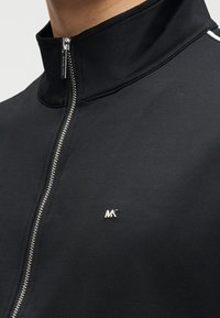 Μαύρο μπουφάν με φερμουάρ και ψηλό γιακά, με ασημένιο φερμουάρ και μικρό μεταλλικό λογότυπο στο στήθος, κατασκευασμένο από απαλό, λείο ύφασμα.