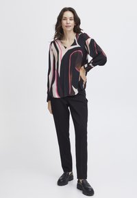 Blouse noire et crème avec des motifs abstraits roses et bruns, dotée d'un décolleté en V et de manches à revers, associée à un pantalon noir et des mocassins.