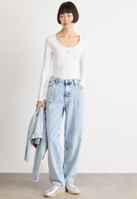 Haut blanc à manches longues avec encolure dégagée, jean taille haute bleu clair, veste en denim portée sur le bras, baskets grises avec semelles beige.