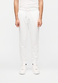 Λευκό βαμβακερό παντελόνι φόρμας με κορδόνι στη μέση, στενά πόδια και πλαϊνές τσέπες, φορεμένο με λευκά αθλητικά παπούτσια. Λείο υλικό, μίνιμαλ σχεδιασ μός.