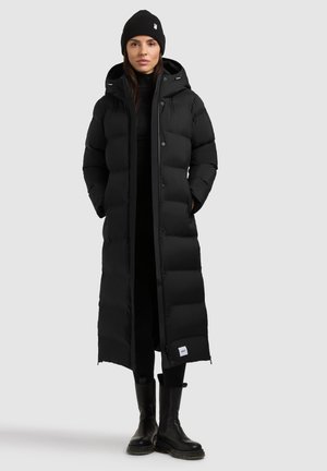 Mujer con un abrigo acolchado largo negro con capucha, gorro negro, jersey de cuello alto negro, pantalones negros y botas negras, de pie frente a un fondo liso.