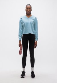 Γυναίκα που στέκεται, φοράει ανοιχτό μπλε φούτερ, μαύρο κολάν, λευκές κάλτσες, μαύρα αθλητικά παπούτσια, κρατώντας ροζ λάστιχο αντίστασης στο δεξί χέρι.