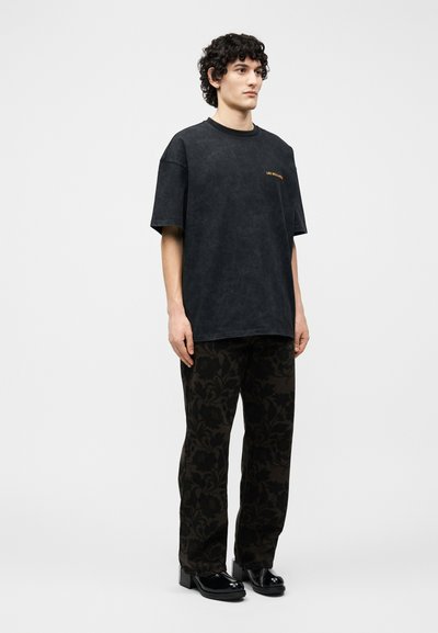 Άνδρας με κατσαρά μαλλιά, φορώντας μια μαύρη φαρδιά μπλούζα, σκούρο παντελόνι με λουλουδάτο σχέδιο και μαύρα λαμπερά παπούτσια με τετράγωνη μύτη, στέκεται μπροστά σε λευκό φόντο.