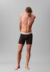 Svarta boxershorts med en kontrasterande beige midjeband från Calvin Klein. Modellen står barfota och visar upp en slank atletisk kroppsbyggnad.