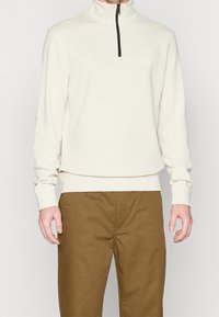 Svetle béžová zipová mikina s vysokým límcem, texturovaná látka, černý zip a malá nášivka s logem na hrudi. V kombinaci s hnědými kalhotami.