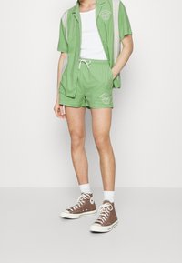Camisa de algodón verde de manga corta con rayas blancas verticales, combinada con pantalones cortos a juego. Zapatillas altas marrones y calcetines blancos completan el look.