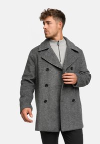 Cappotto doppiopetto in misto lana grigia con grandi reverse e bottoni neri, indossato sopra un maglione grigio chiaro a collo alto.