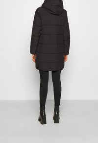 Abrigo acolchado negro con capucha, que presenta un diseño de costura horizontal, mangas largas y una longitud hasta la mitad del muslo. Combinado con leggings negros y botines.