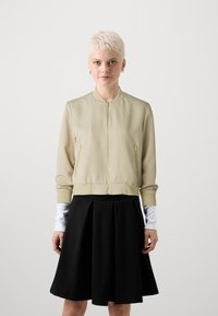 Světle béžová bomber bunda se zipem vpředu, bočními kapsami, žebrovanými manžetami a lemem, v kombinaci s černou rozšířenou sukní. Hladká textura.