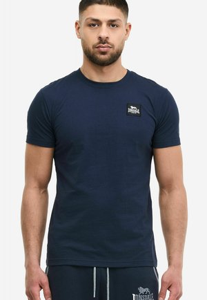 Muži v přiléhavém tmavě modrém tričku s malou nášivkou s logem Lonsdale London na levé straně hrudi, kteří stojí před jednobarevným pozadím.