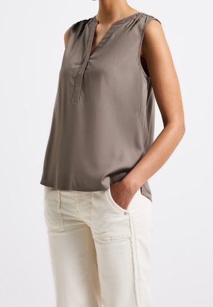 Blusa sin mangas color topo con escote en V, llevada sin meter por dentro sobre pantalones beige claros, con una mano casualmente en el bolsillo sobre un fondo liso.