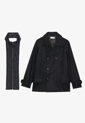 Zwarte wollen peacoat met dubbele knopen en grote kraag, gecombineerd met een mouwloze zwarte gewatteerde vest met ritssluiting.